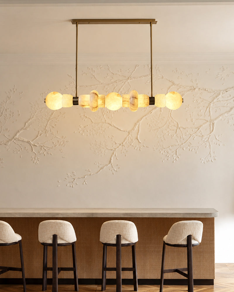 A modern multi-light linear pendant chandelier with warm glowing glass globes and a brass-toned frame, hanging above a bar counter with four stools in a styled interior setting.