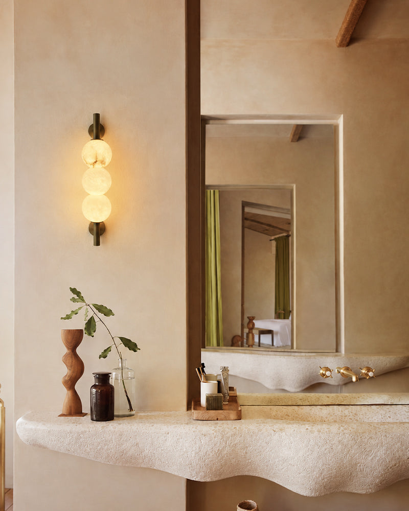 A modern wall-mounted light fixture with three stacked frosted glass spheres and a dark metal frame, glowing warmly on a beige plaster bathroom wall beside a large mirror and stone vanity.