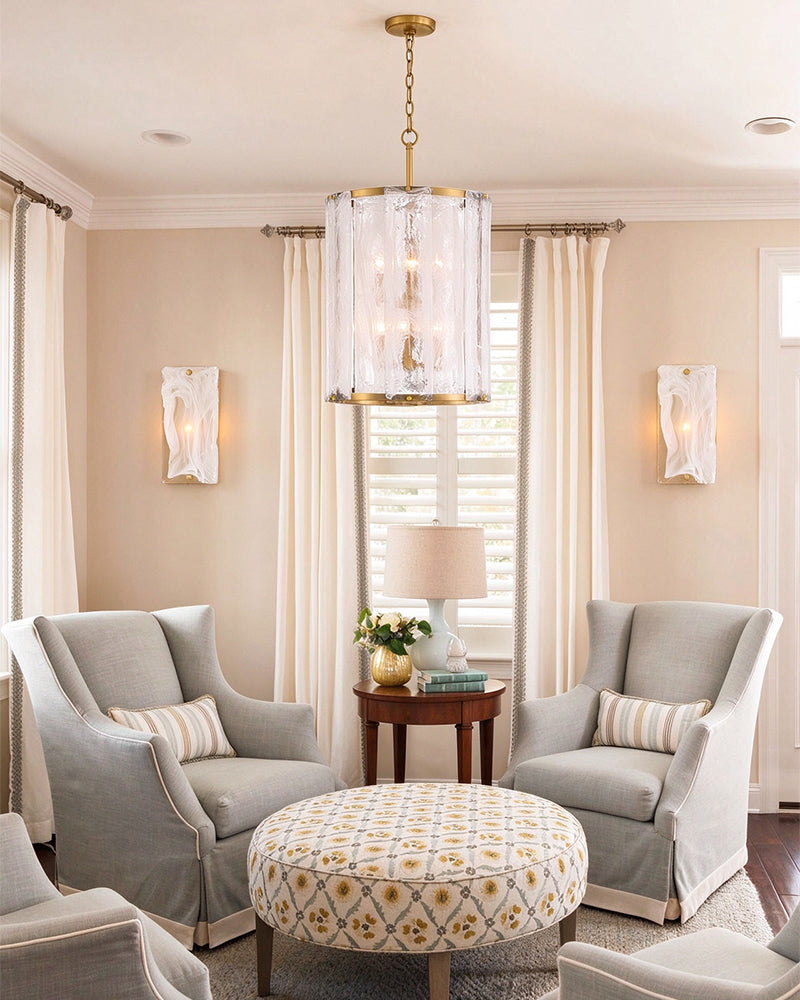 A styled living room featuring a gold and crystal cylinder chandelier hanging above a patterned round ottoman. The room is furnished with two light gray slipcovered armchairs, a small wooden side table with a lamp, and draped cream curtains against beige walls.