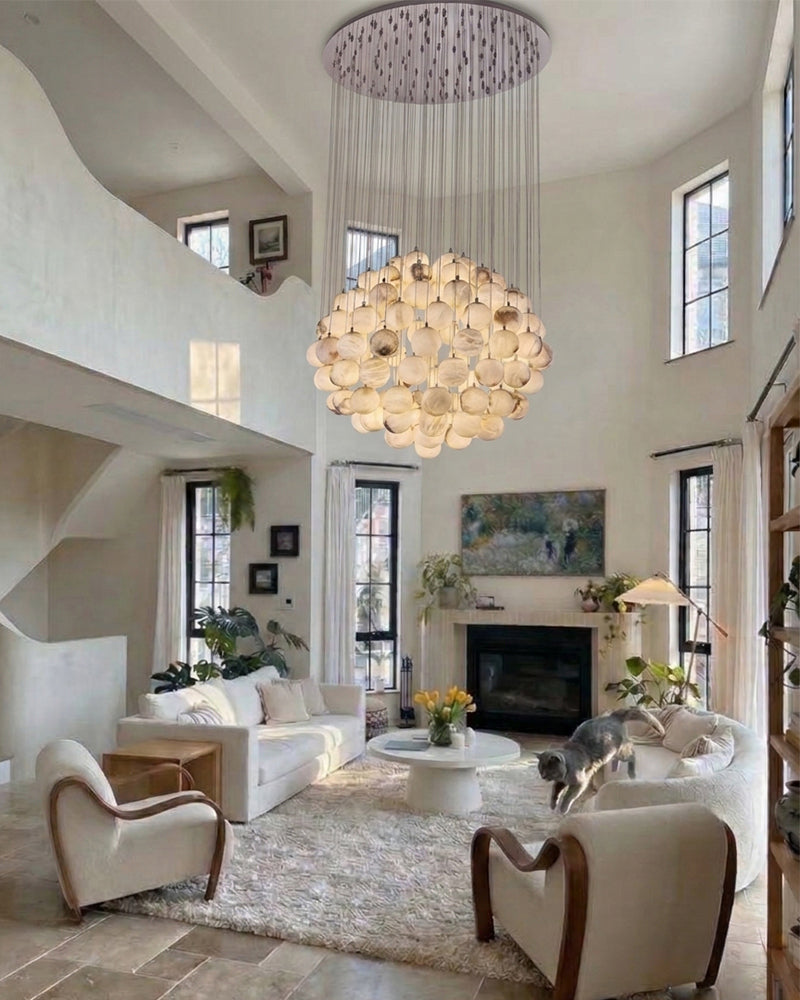 A large, modern multi-light chandelier with many curved glass shades hangs in a bright, high-ceilinged living room with cream sofas, a fireplace, and a dog on a rug.