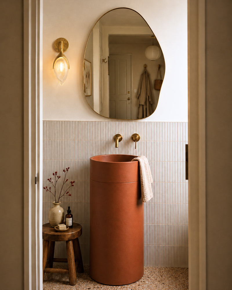 A modern bathroom scene featuring a tall, cylindrical terracotta-colored pedestal sink. The sink is set against a wall with a white upper section and light beige vertical tile lower section, with two matching brass wall-mounted faucets.