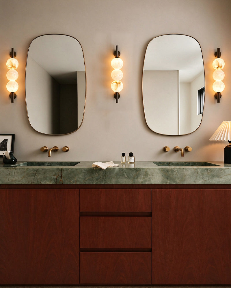 A modern bathroom vanity featuring a dark green marble countertop, a reddish-brown wooden cabinet, two large rounded rectangular mirrors, and three glowing bubble-style wall sconces.