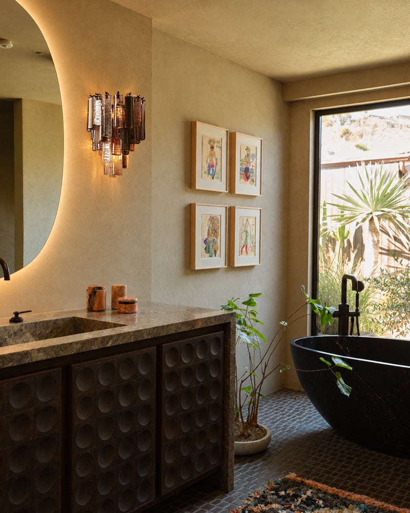 A luxury bathroom interior featuring a dark wood vanity with a marble countertop, a round black freestanding bathtub by a large window, and warm beige walls with framed art and a sculptural wall sconce.