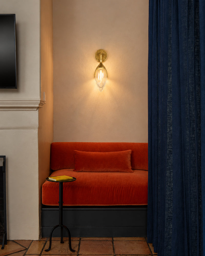A warm brass wall sconce with a clear glass shade mounted on a beige wall above an orange velvet bench with a matching pillow, next to a dark blue curtain.