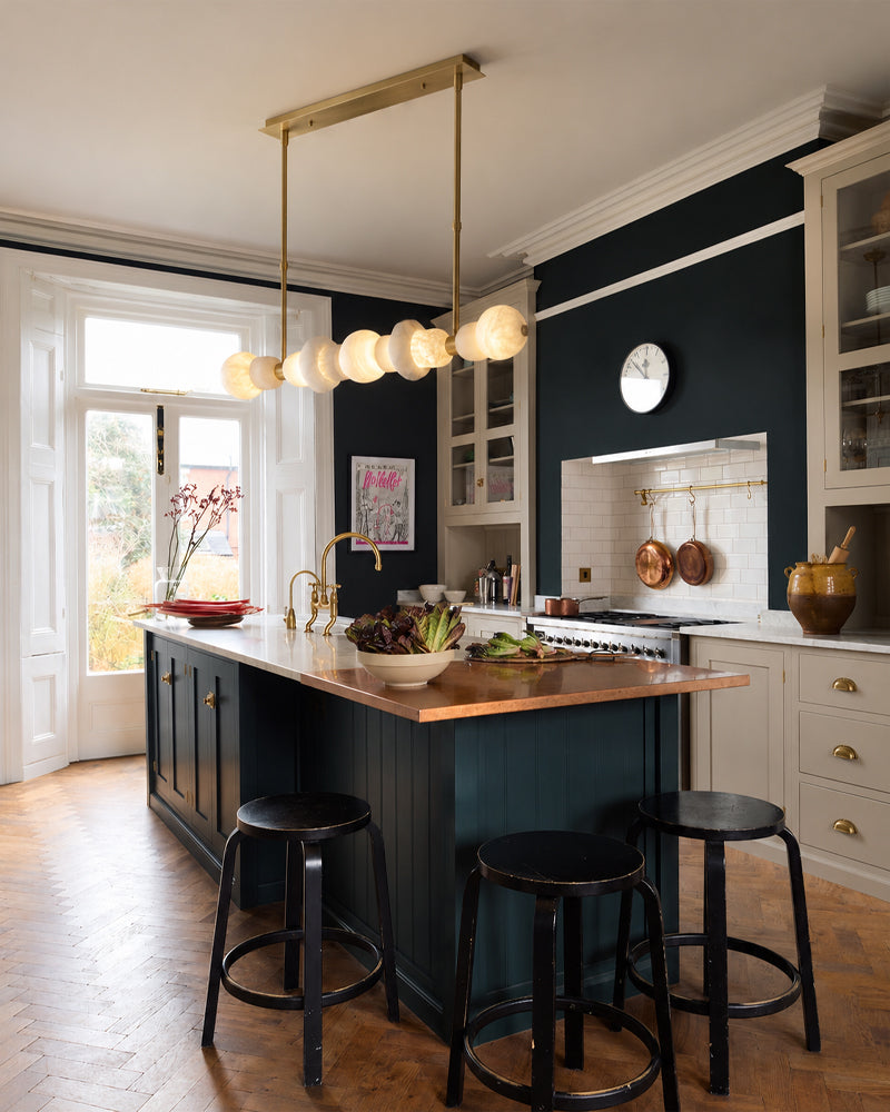 A modern kitchen interior with a dark navy blue island, light wood countertops, black bar stools, and a brass multi-light pendant fixture. White cabinets, a tiled backsplash, and wood herringbone flooring are visible in the room.