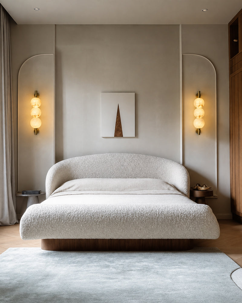 A modern bedroom featuring a low-profile bed with a textured boucle upholstered headboard and matching bedding. The room has warm neutral walls with arched recesses housing glowing three-globe wall sconces, a minimalist canvas print, and a light blue-gray rug on a wood floor.