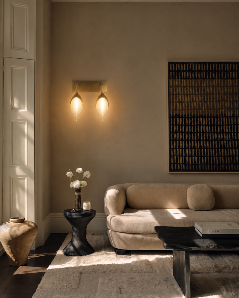 A warm, modern living room scene featuring a beige sofa, a dual-light wall sconce with glowing bulbs, a black side table with white flowers, and a textured dark wall art piece.