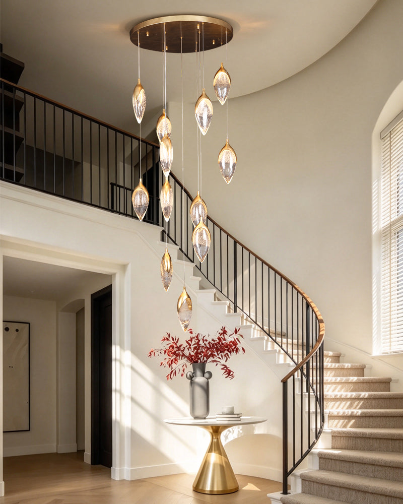 A modern multi-light pendant chandelier with clear teardrop glass drops and brushed gold-toned metal accents, hanging in a bright, elegant staircase hall with beige walls and a curved black metal railing.