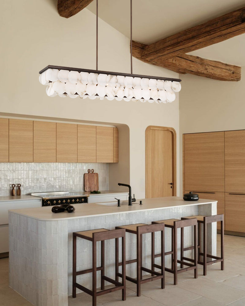 A modern kitchen interior with a long rectangular pendant light featuring many small white globe bulbs suspended above a light stone island. The room has exposed wooden ceiling beams, light wood cabinetry, and dark wood bar stools at the island.