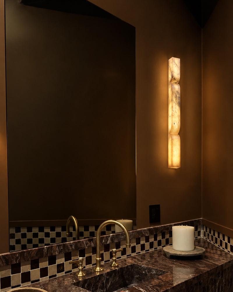 A modern vertical wall sconce made of carved natural stone, glowing with a warm amber light, mounted on a dark brown bathroom wall above a marble vanity with brass fixtures.