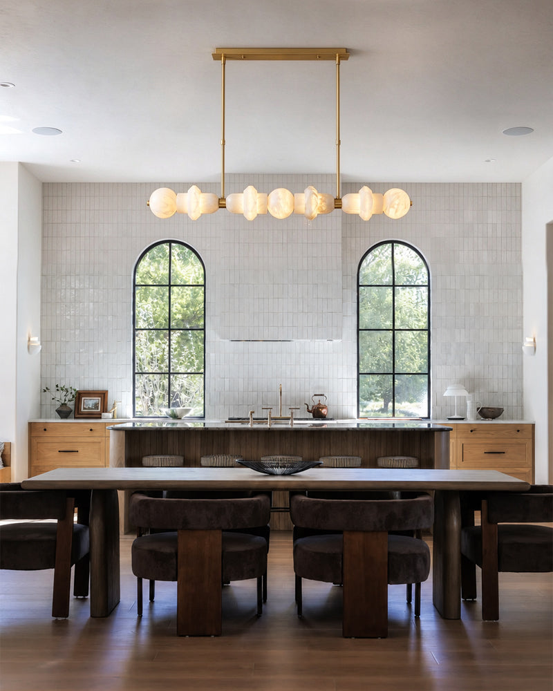 A modern dining room with a long rectangular wooden table and six dark upholstered chairs, set in a bright kitchen with white tile walls, arched windows, and a gold-framed chandelier overhead.