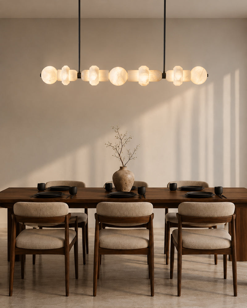 A modern dining room scene with a long wooden table, six upholstered chairs, a ceramic vase with dried branches, and a large multi-light linear pendant chandelier casting warm shadows on the wall.