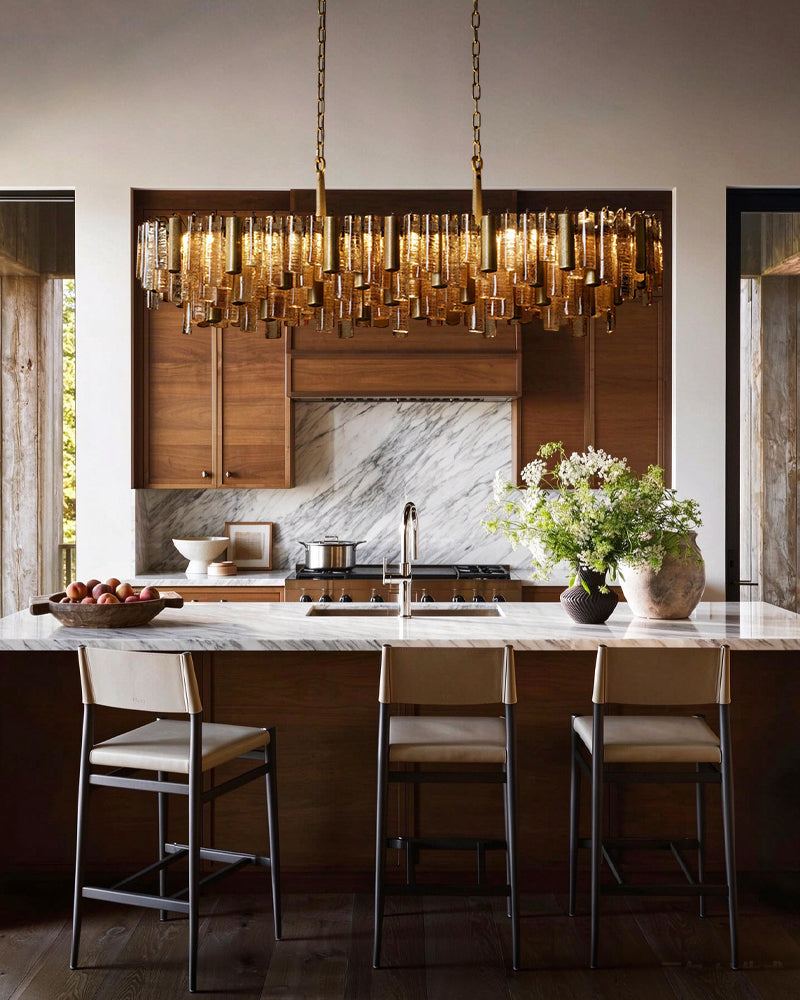 A modern kitchen interior with a large gold-toned chandelier hanging above a marble island, wood cabinetry, three bar stools, and a vase of white flowers on the counter.