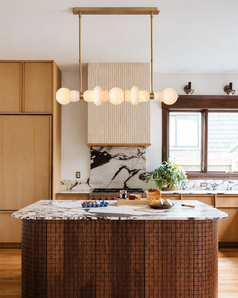 A modern kitchen interior with a large island featuring a brown wood-grain tiled base and a white marble countertop. A brass pendant light with multiple frosted globe bulbs hangs above the island.