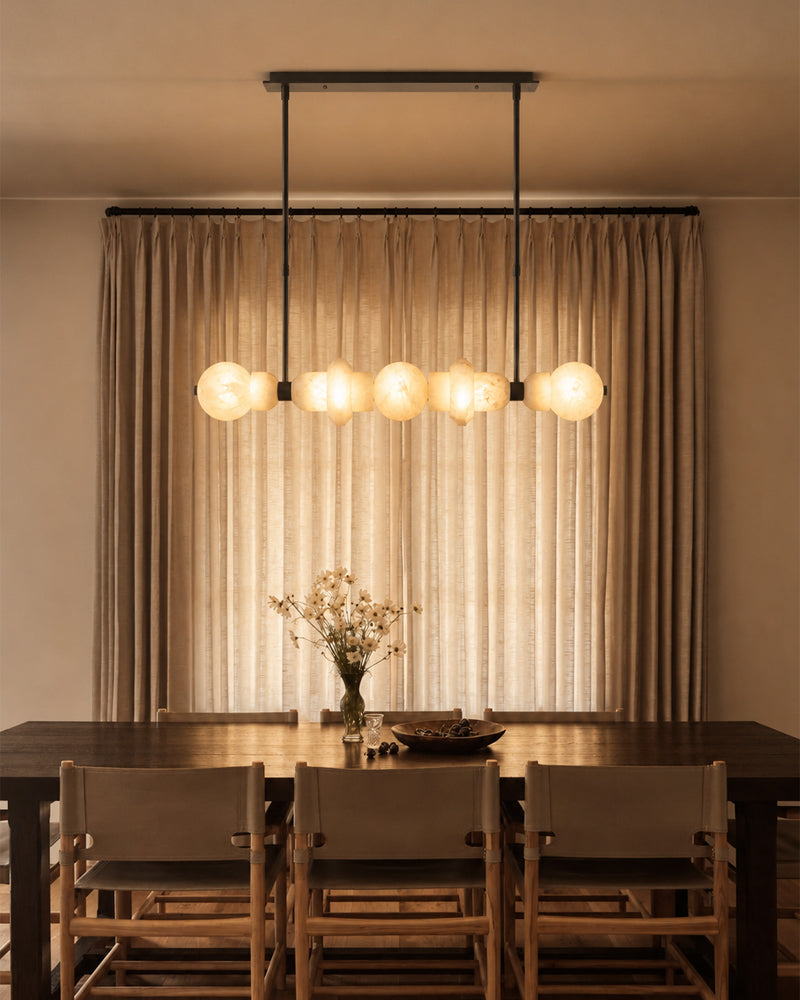 A modern multi-light linear pendant chandelier with six glowing spherical glass shades hangs over a dining table with chairs in a warm, neutral-toned dining room.
