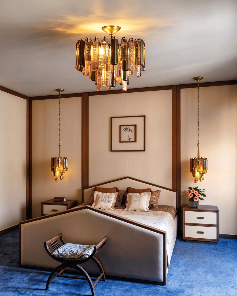 A luxury bedroom featuring a modern bed with a low upholstered headboard and footboard, flanked by matching nightstands. The room is decorated with a blue carpet, warm wood trim, dual hanging pendant lights, and a prominent gold-toned cylindrical chandelier on the ceiling.