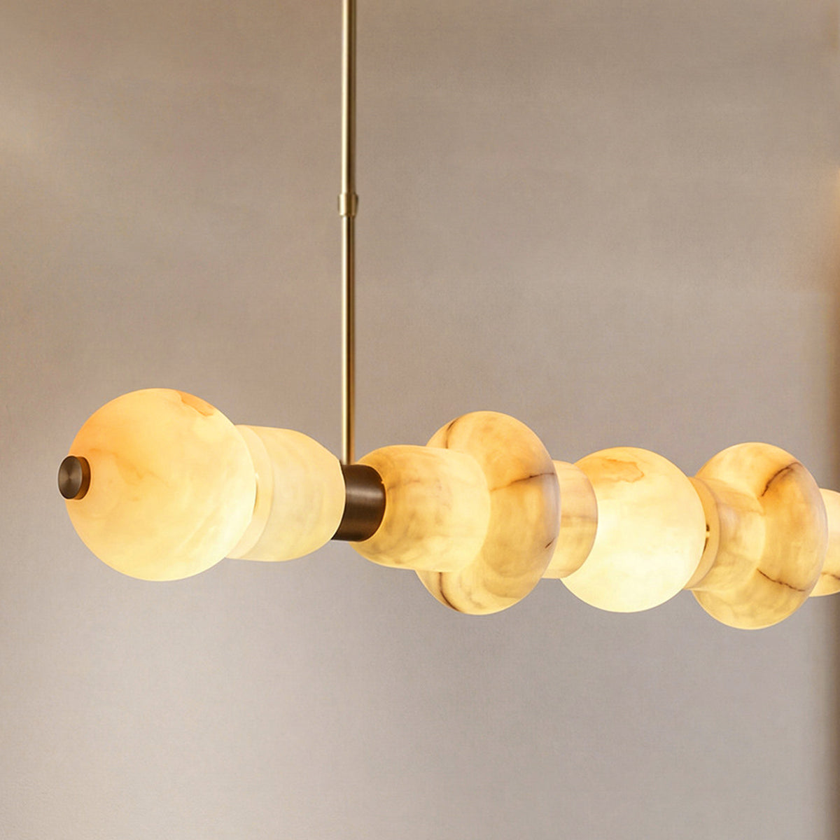 A modern linear pendant light with a series of organic, rounded amber-colored glass or acrylic beads suspended by a slim brass rod against a neutral gray background.