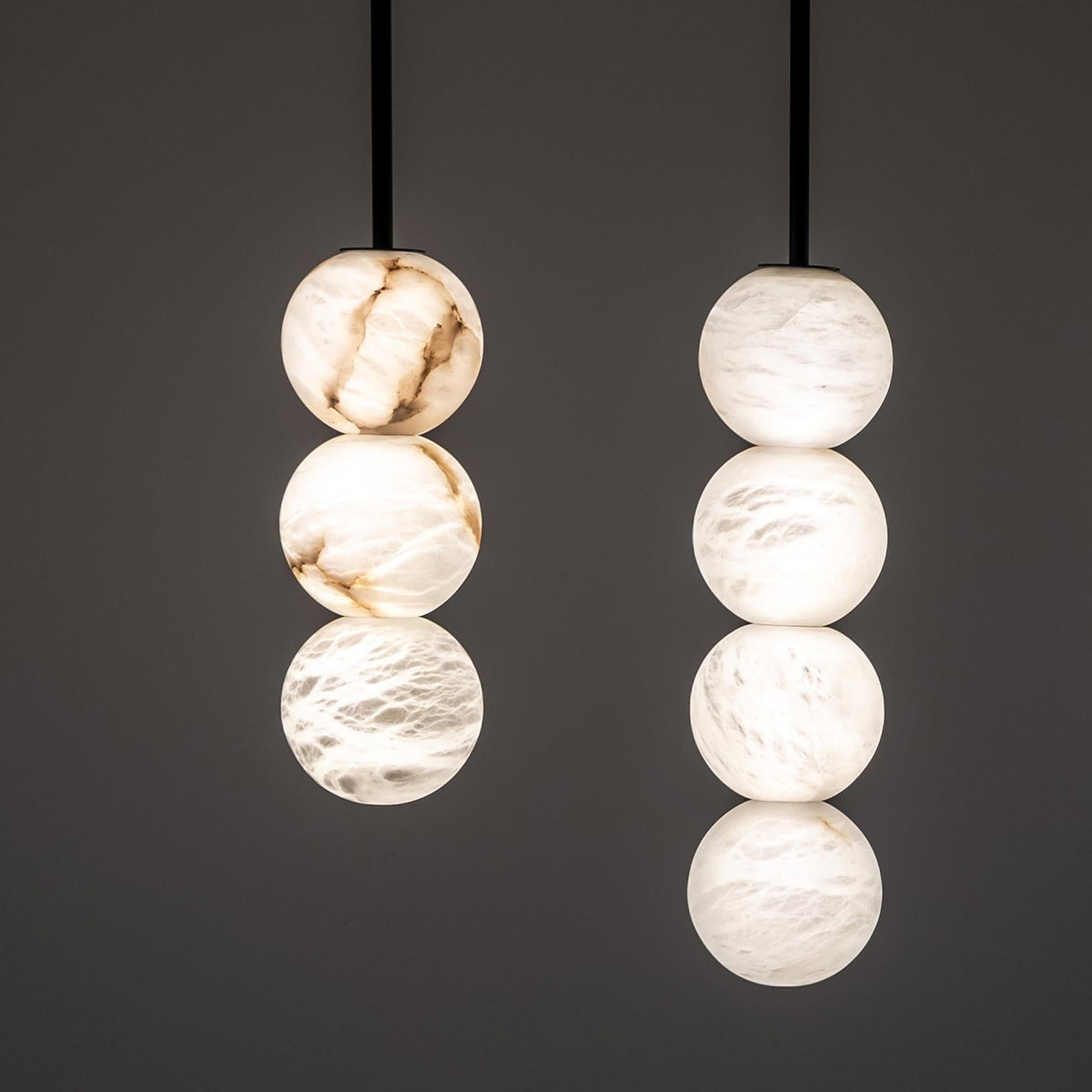 Two modern hanging pendant lights with stacked spherical glass shades, one in polished white onyx and one in frosted white glass, suspended against a dark gray background.