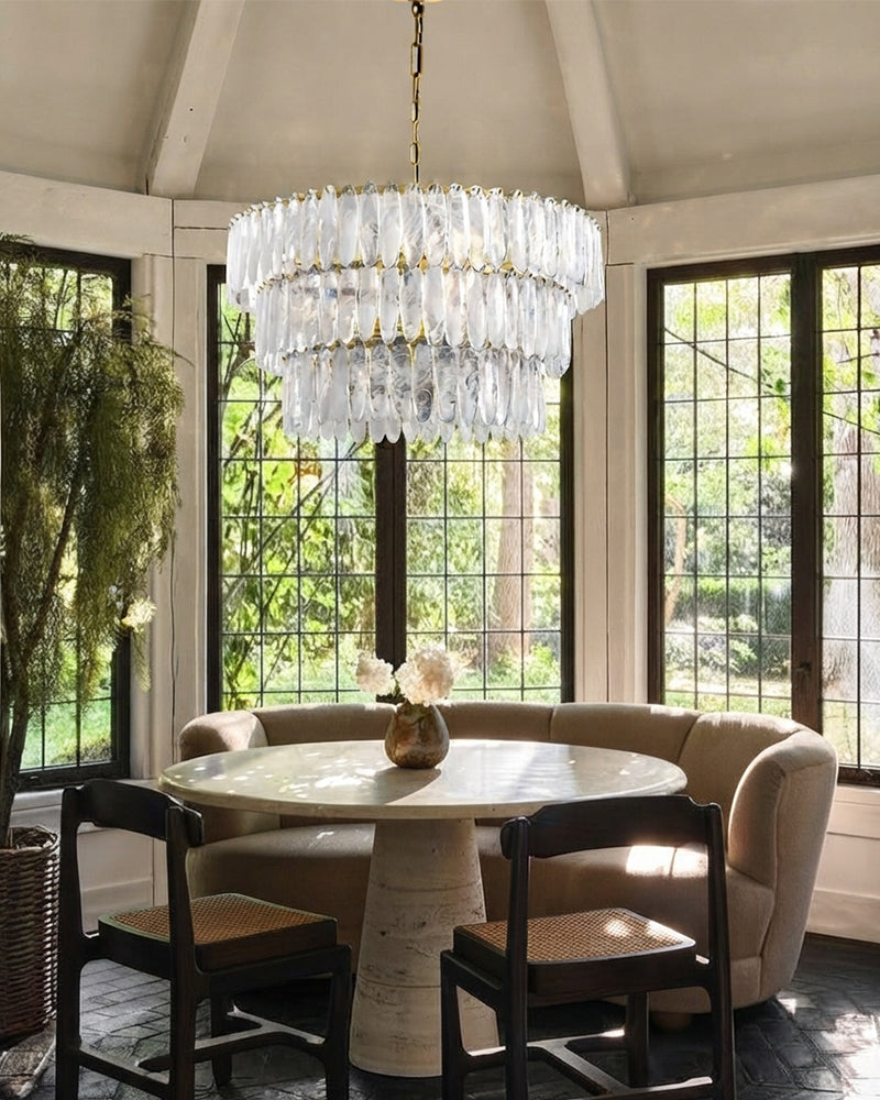 A large tiered glass chandelier with gold-toned hardware hangs over a round dining table in a sunlit room with a curved upholstered bench, dark wood chairs, and tall windows looking out to greenery.