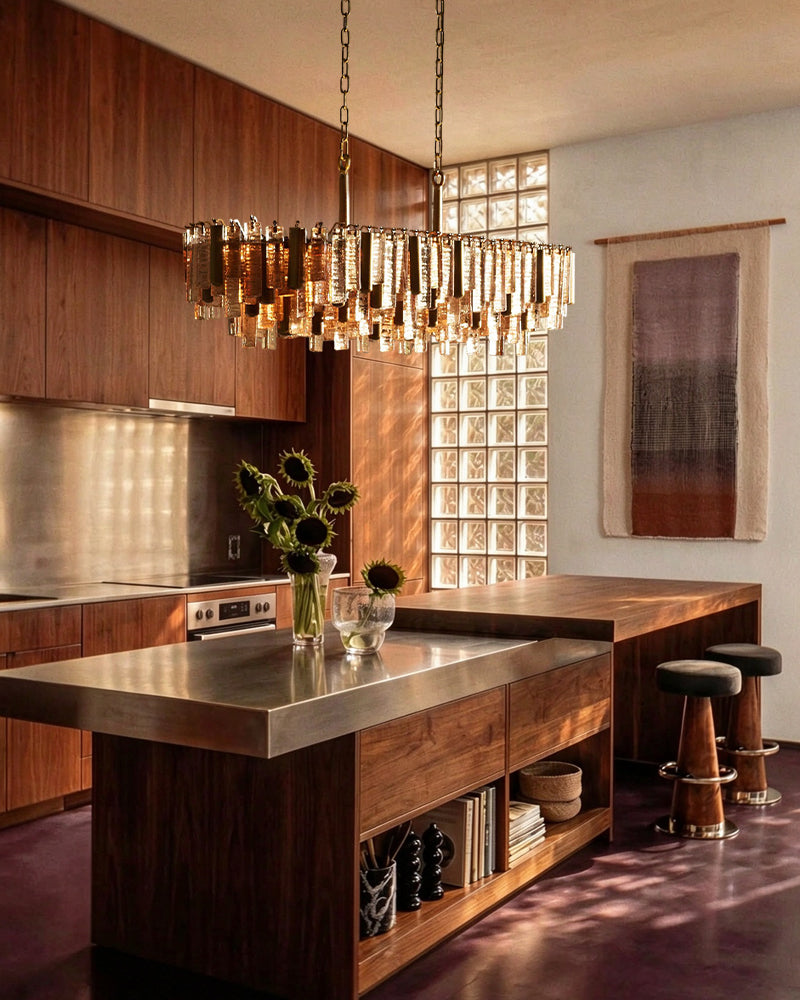 A modern kitchen interior with dark wood cabinetry and island, a large hanging chandelier with clear cylindrical glass drops, a glass block window, two bar stools, and warm sunlight streaming across the space.