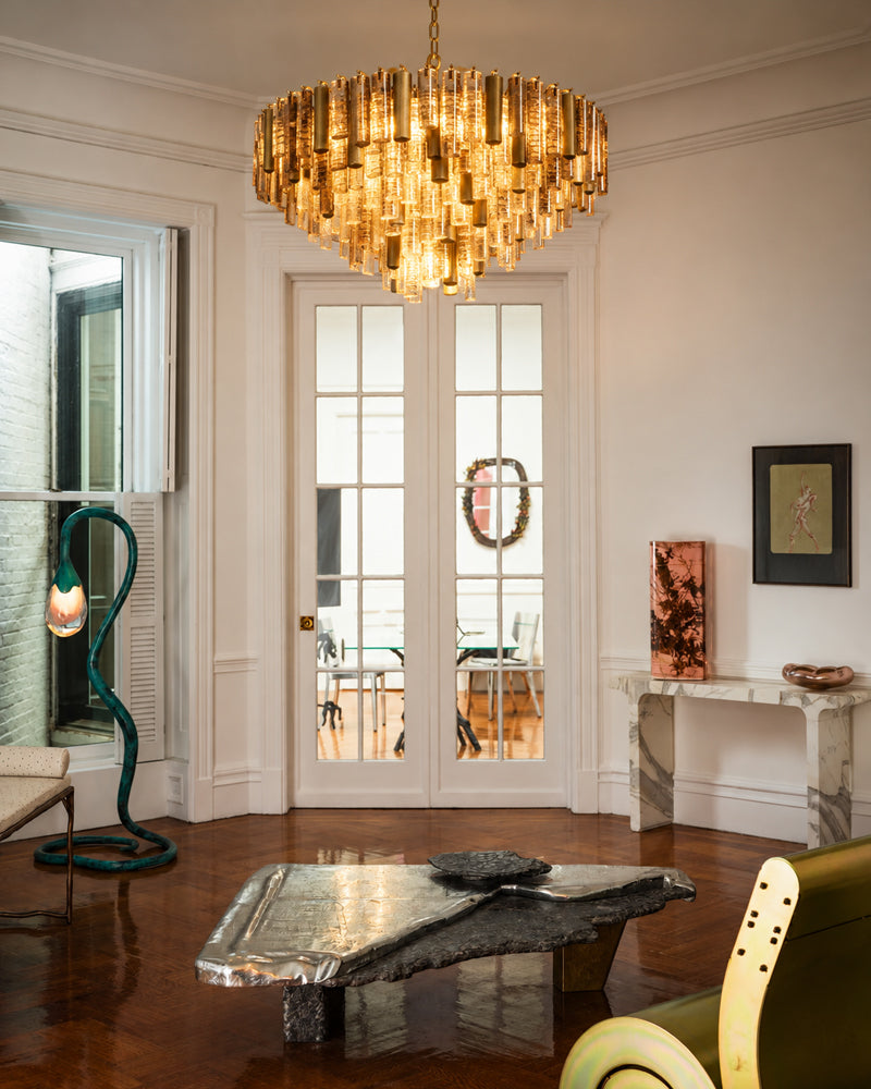 A large cylindrical chandelier with a cascading gold-toned glass rod design hangs in a bright, elegant living room with white walls, double glass doors, and a polished wood floor.