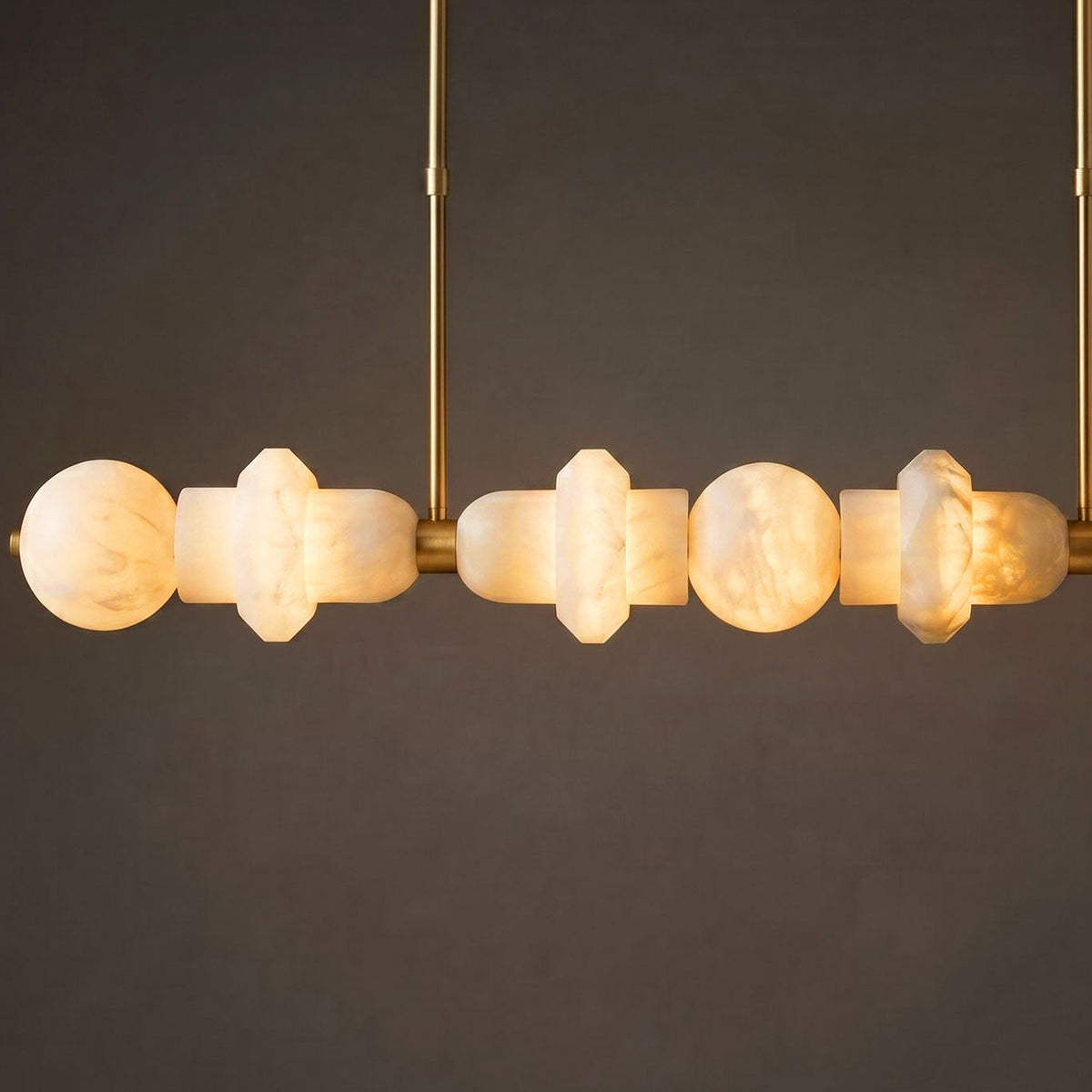 A modern multi-light linear pendant chandelier with warm white marble beads and a brushed brass finish, suspended against a dark gray background.
