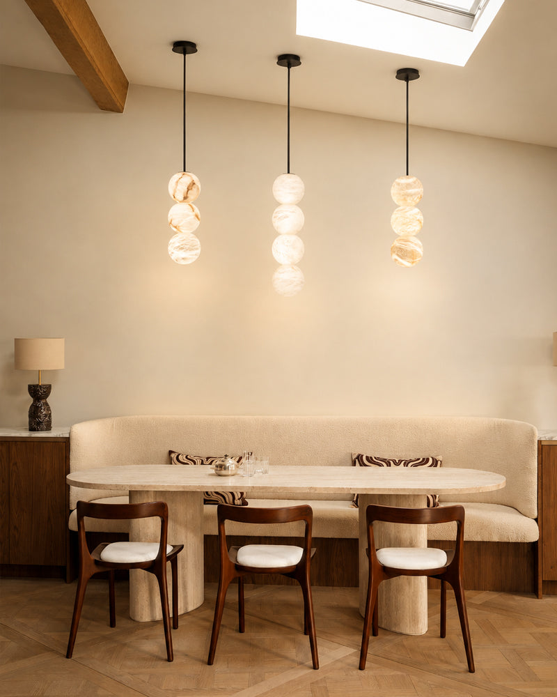 A modern dining room featuring three hanging pendant lights with stacked glass globes, a cream upholstered banquette, a light stone dining table, and four wooden chairs with white seat cushions.