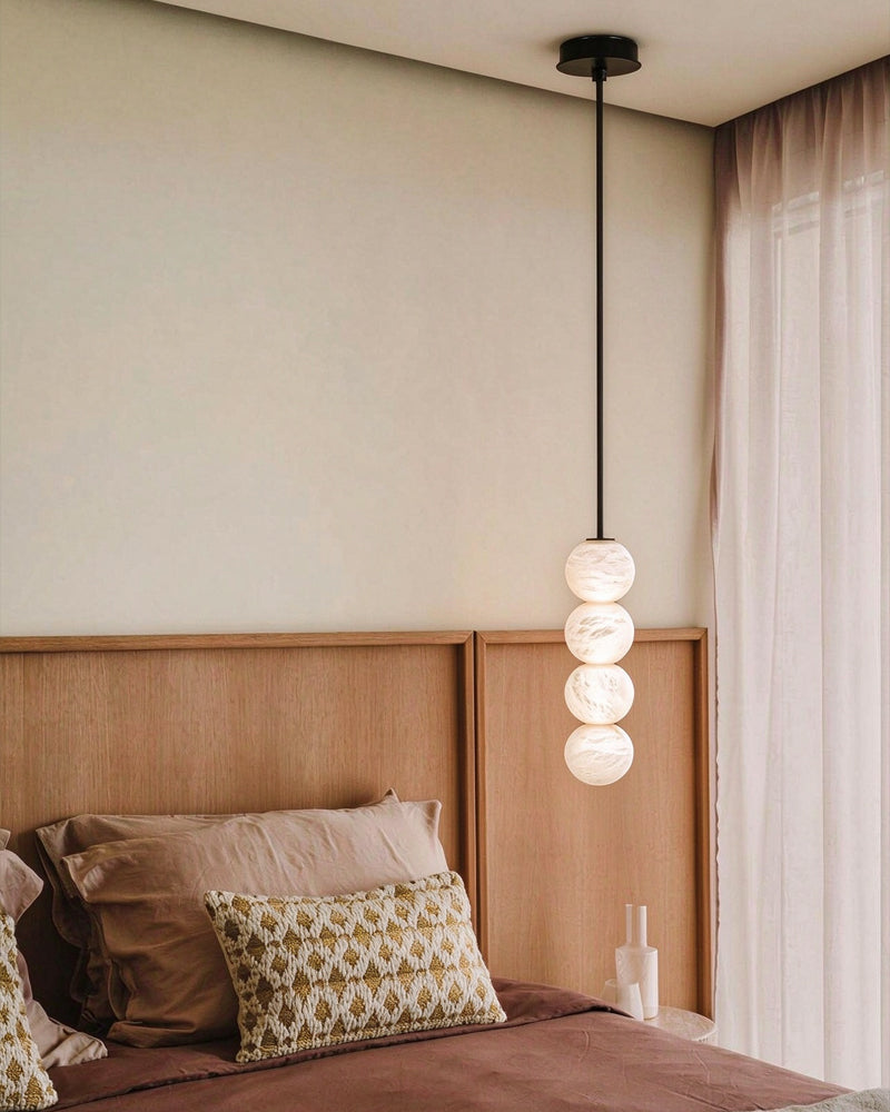 A modern pendant light with four stacked spherical glass shades hangs above a bed with a wooden headboard in a softly lit bedroom.