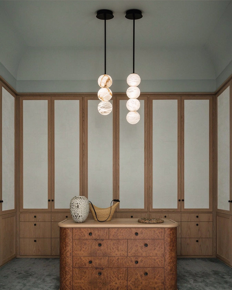 Two modern pendant light fixtures hang above a wooden chest in a styled interior. Each fixture has a black cord and canopy, with one cluster of three white marble-like spherical shades and the other of four white marble-like spherical shades.
