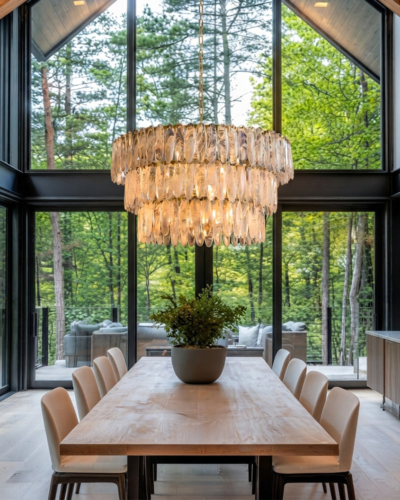 A modern tiered glass chandelier hangs over a light wood dining table with beige upholstered chairs in a bright dining room with large black-framed windows overlooking a forest.