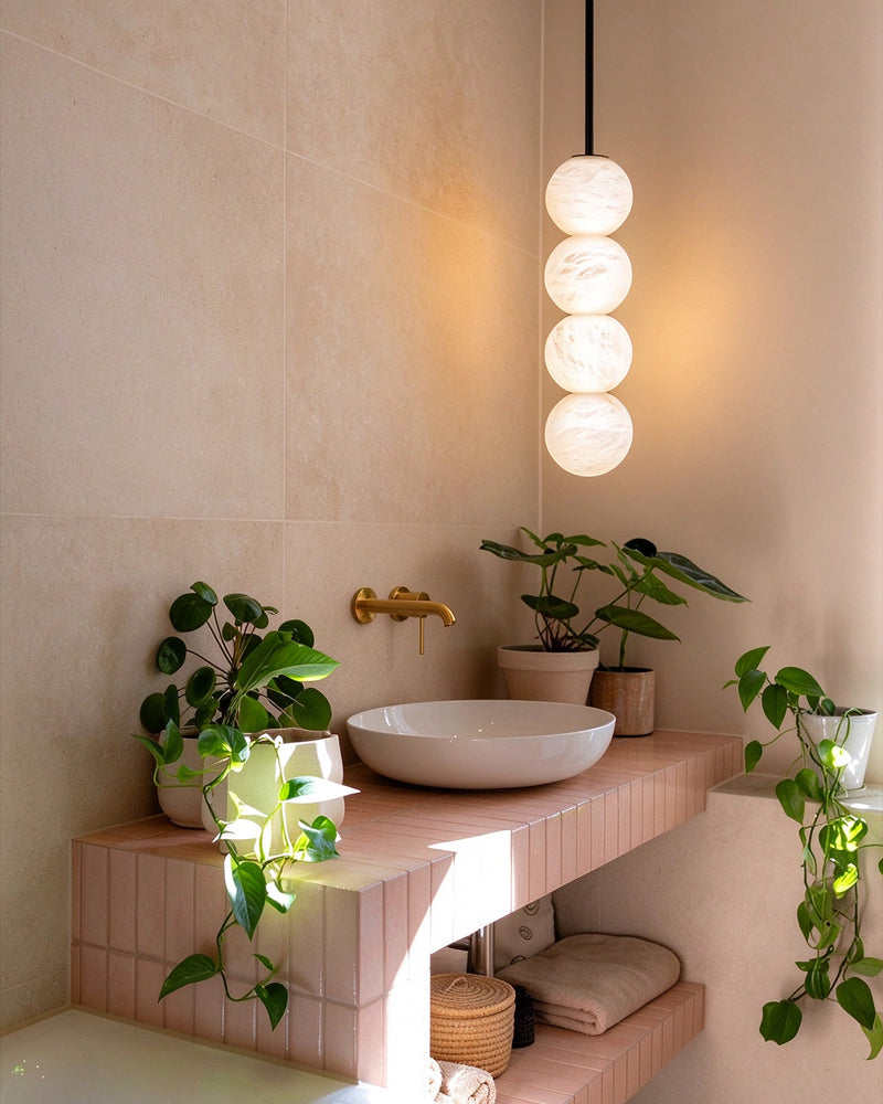 A modern bathroom vanity setup with a white oval sink, pink tiled counter, gold faucet, hanging multi-sphere pendant light, and several green potted plants in a warm beige room.