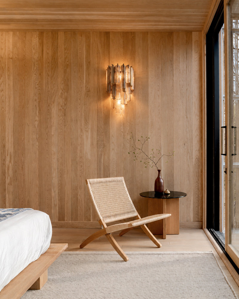 A modern wooden wall sconce with vertical glass shades is mounted on a wood-paneled bedroom wall above a woven accent chair and a small side table.
