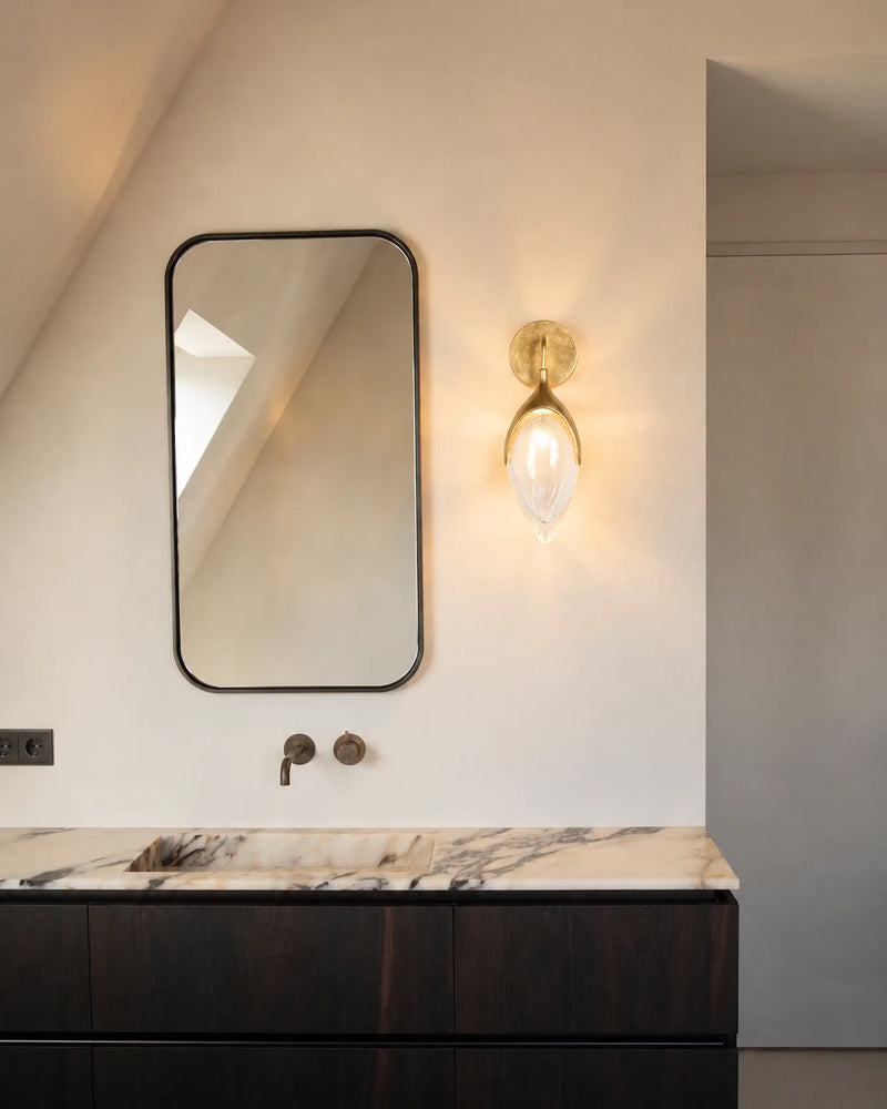 A modern bathroom vanity featuring a rectangular wall mirror with a thin black frame, a warm brass wall sconce, and a dark wood cabinet with a marble countertop.