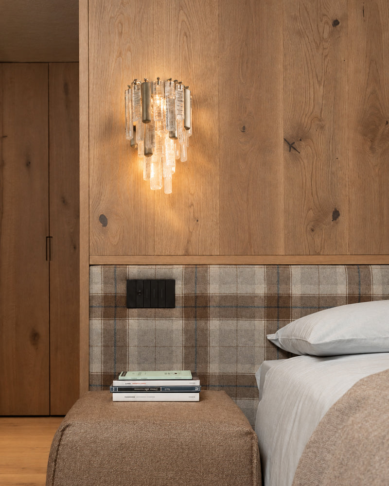 A modern bedroom scene featuring a wall-mounted glass chandelier with a warm glowing light, a wood-paneled wall, a plaid headboard, and a bed with white linens.