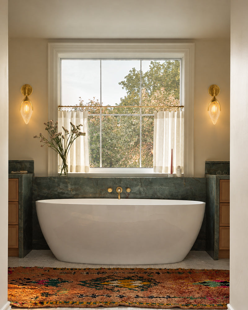 A freestanding oval bathtub in a styled bathroom setting, positioned in front of a dark stone wall and a window with sheer curtains and outdoor greenery. The room features warm beige walls, brass sconces, a patterned rug, and wooden vanity cabinets.