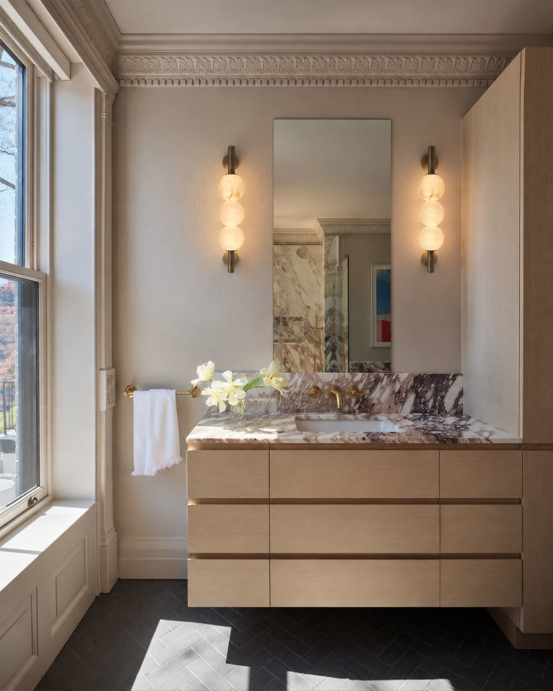 A modern bathroom vanity featuring a light wood floating cabinet, a white and brown marble countertop with an integrated sink, a large rectangular mirror, and two matching wall sconces. The room has beige walls, a window on the left with a white towel hanging from its rail, and dark herringbone tile flooring.