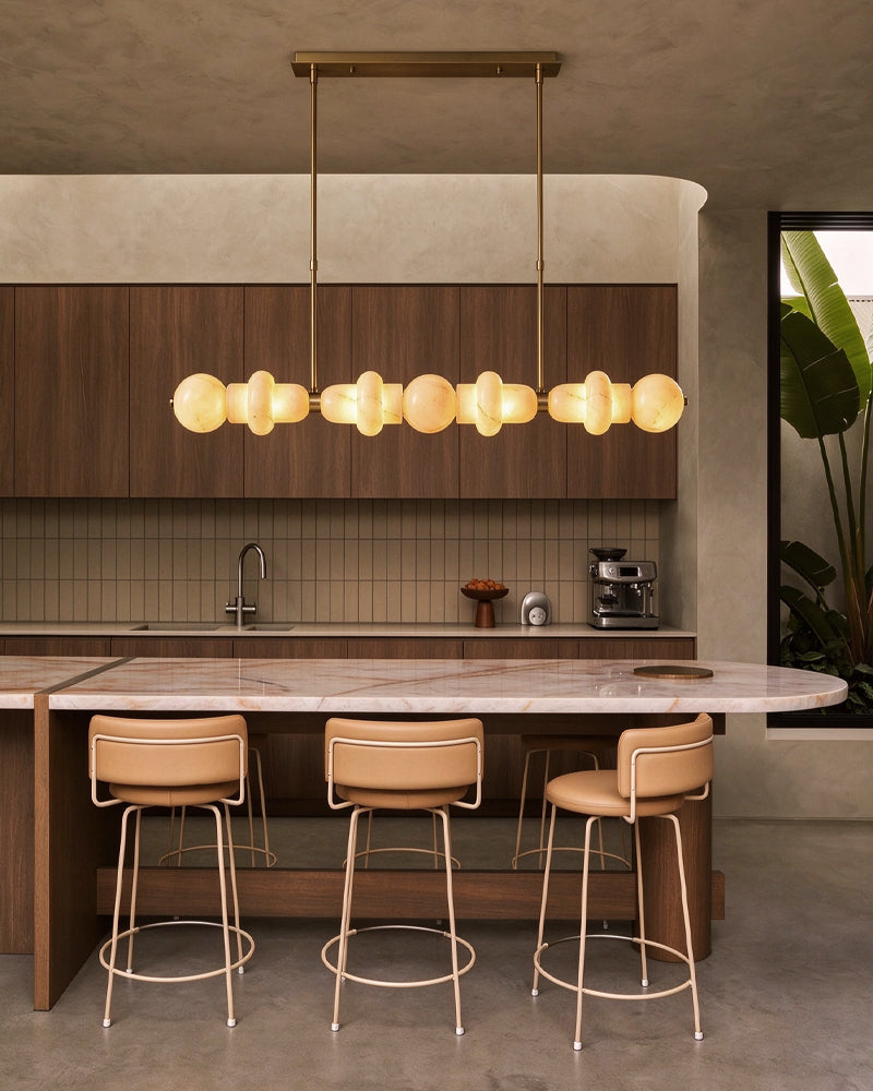 A modern kitchen interior with a long linear pendant light featuring multiple warm glowing frosted globe bulbs suspended above a marble-topped island. Three tan leather bar stools with gold metal frames sit beneath the light.