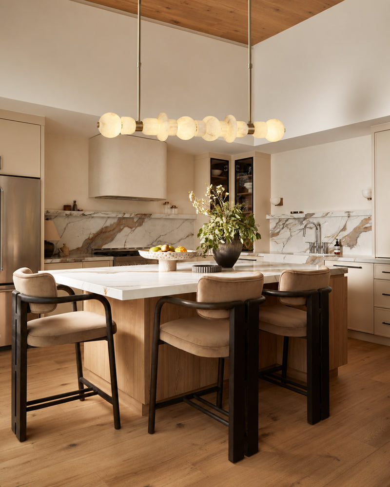A modern kitchen island with a white marble countertop and wood base, surrounded by four upholstered bar stools with dark wood frames. The kitchen features cream cabinetry, a marble backsplash, a wood ceiling, and a multi-light pendant fixture.
