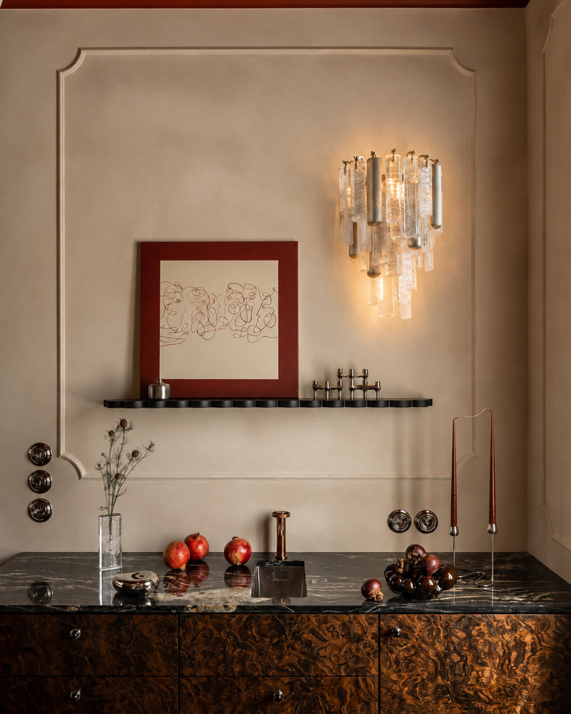 A luxury interior scene featuring a modern wall-mounted chandelier with clear glass cylindrical tubes and warm gold-toned metal accents, illuminated against a beige wall. Below, a dark wood cabinet with a black marble countertop holds decorative objects, with a red-framed print on a black shelf above.