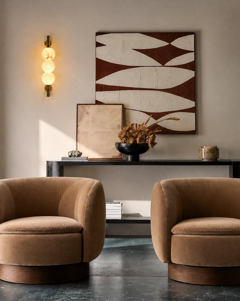 A styled interior scene with two tan upholstered barrel chairs facing a console table, beneath a large abstract wall artwork. A warm spherical wall sconce glows on the left, and the floor is dark polished stone.
