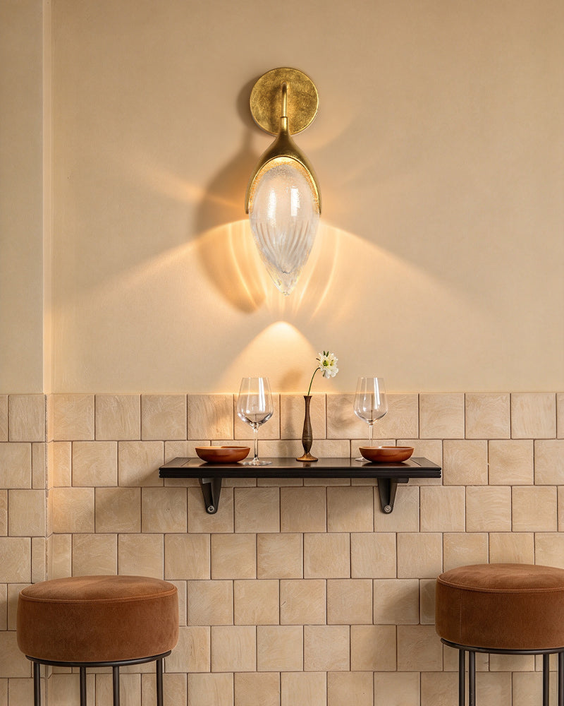 A wall-mounted sconce with a gold-toned circular backplate and a ribbed clear glass shade, casting a warm glow on a beige wall above a shelf with two wine glasses and two brown velvet bar stools.