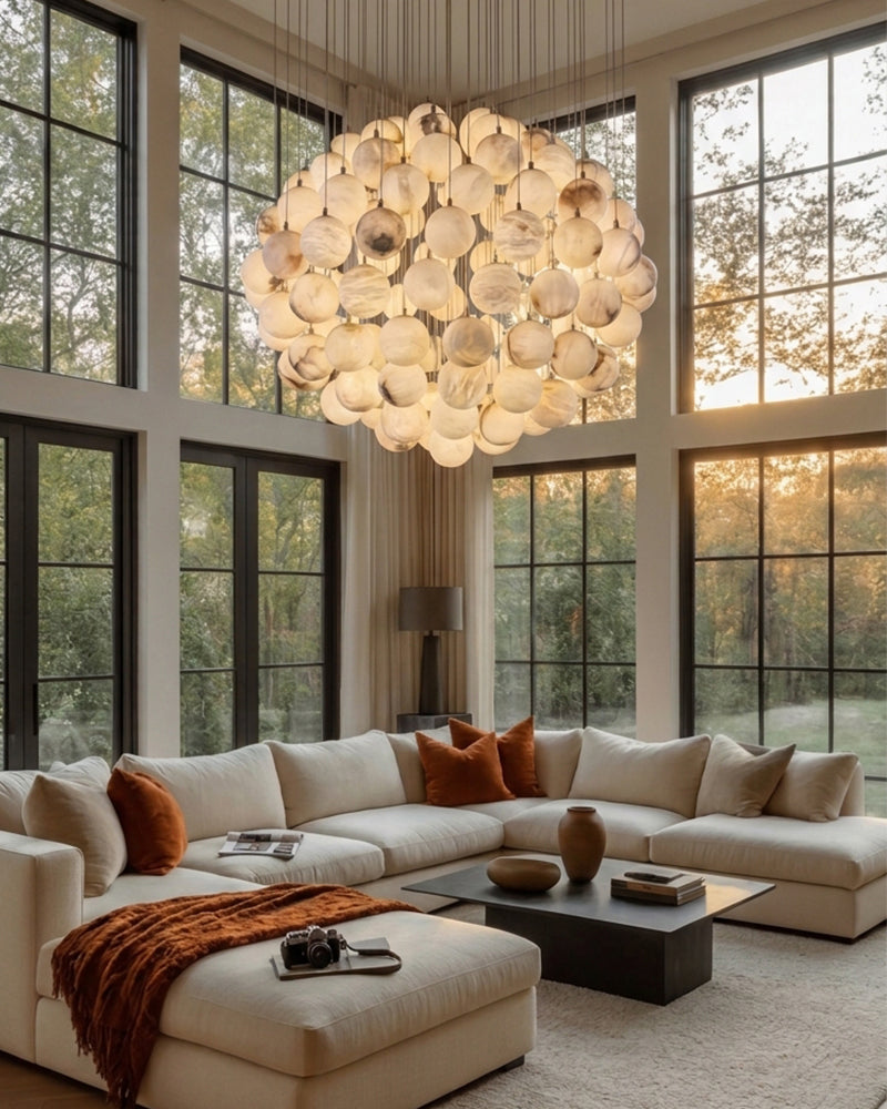 A large, modern multi-light chandelier with many circular glass shades hangs in a bright, high-ceilinged living room with a cream sectional sofa, large black-framed windows, and warm natural light.