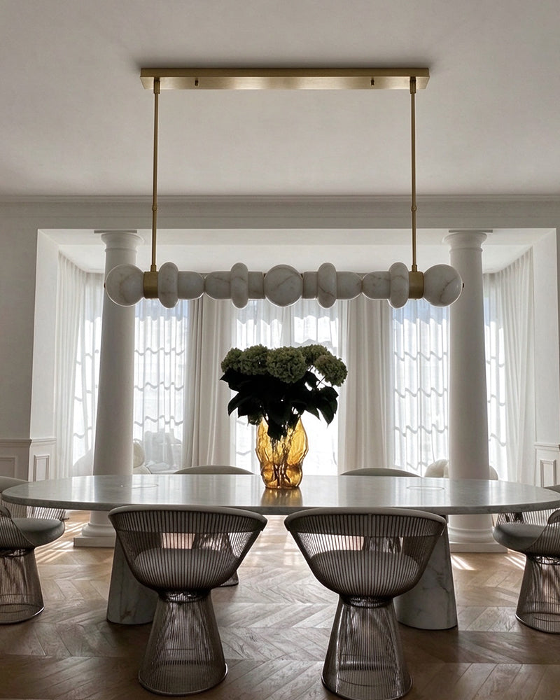 A modern dining room scene featuring a long horizontal chandelier with white spherical glass shades and gold-toned hardware, suspended over a round dining table with wire chairs in a bright interior with white columns and curtains.