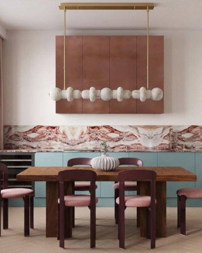 A modern dining room scene featuring a rectangular wooden dining table with six dark wood chairs that have pink cushions, a decorative white ribbed vase, and a large brass pendant light with white sculptural beads hanging above a kitchen area with teal cabinets and a marble backsplash.