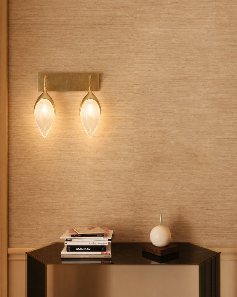 A modern wall-mounted sconce with two teardrop-shaped frosted glass shades and a brushed brass rectangular backplate, shown lit on a beige textured wall above a dark console table with books and a decorative object.