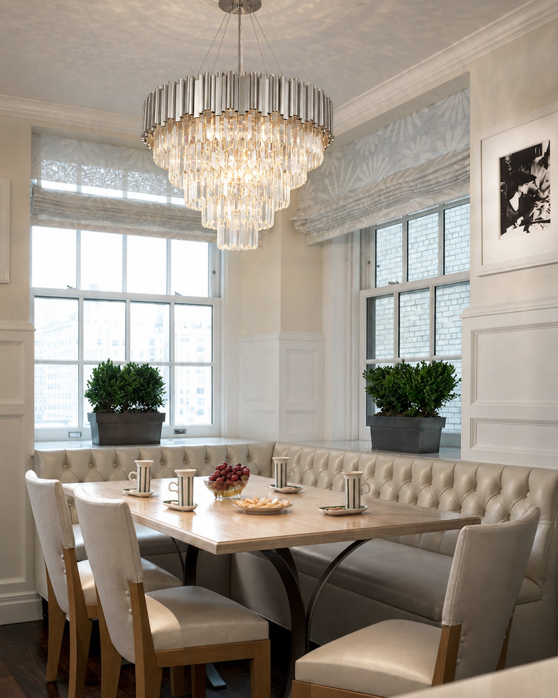 A luxury tiered crystal chandelier hangs over a dining nook with tufted cream banquette seating, a light wood table, and upholstered chairs in a bright, classic dining room.