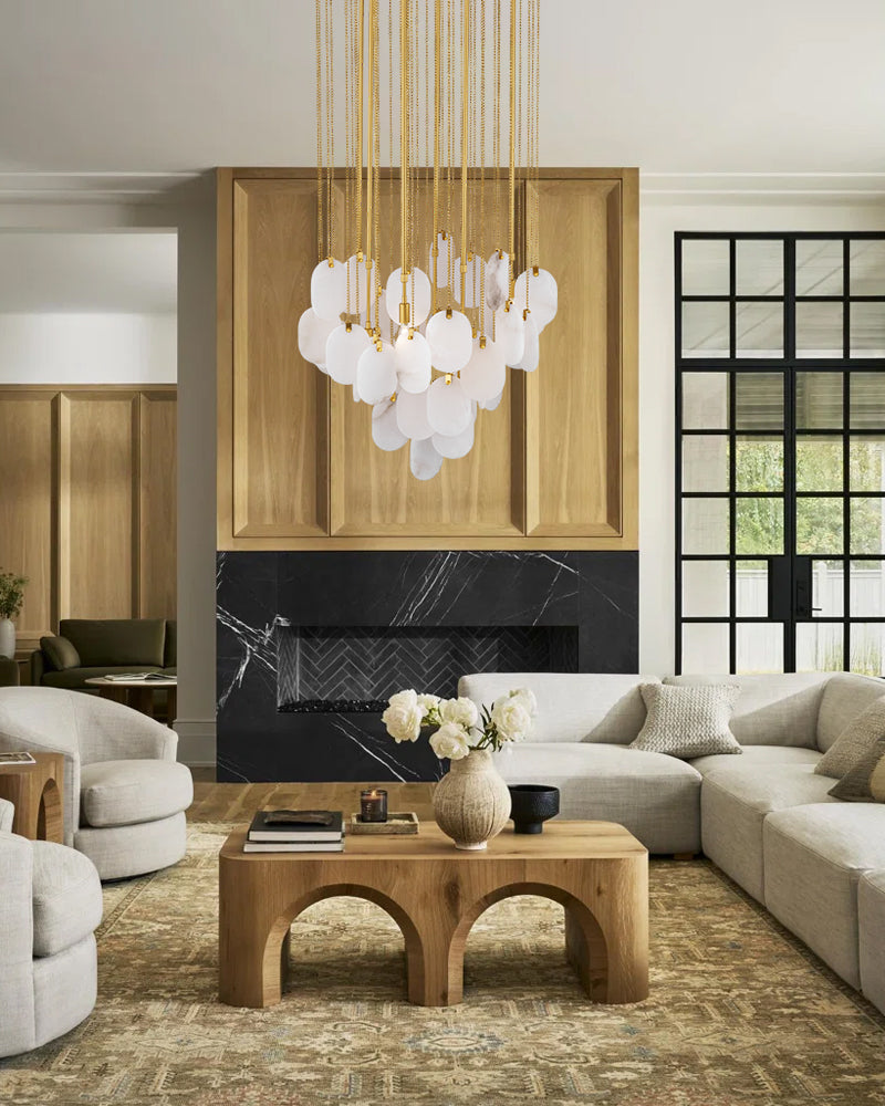 A modern living room featuring a large white and gold bubble chandelier hanging above a black marble fireplace. The room is styled with light beige sectional sofas, a matching wooden coffee table, and a textured area rug.