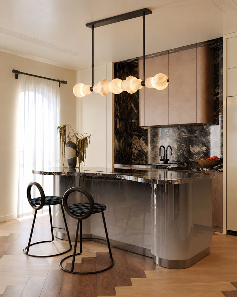 A modern kitchen island with a glossy curved base, dark marble countertop, and two black metal bar stools. A multi-light pendant chandelier hangs above, set in a bright kitchen with wood flooring and dark marble backsplash.