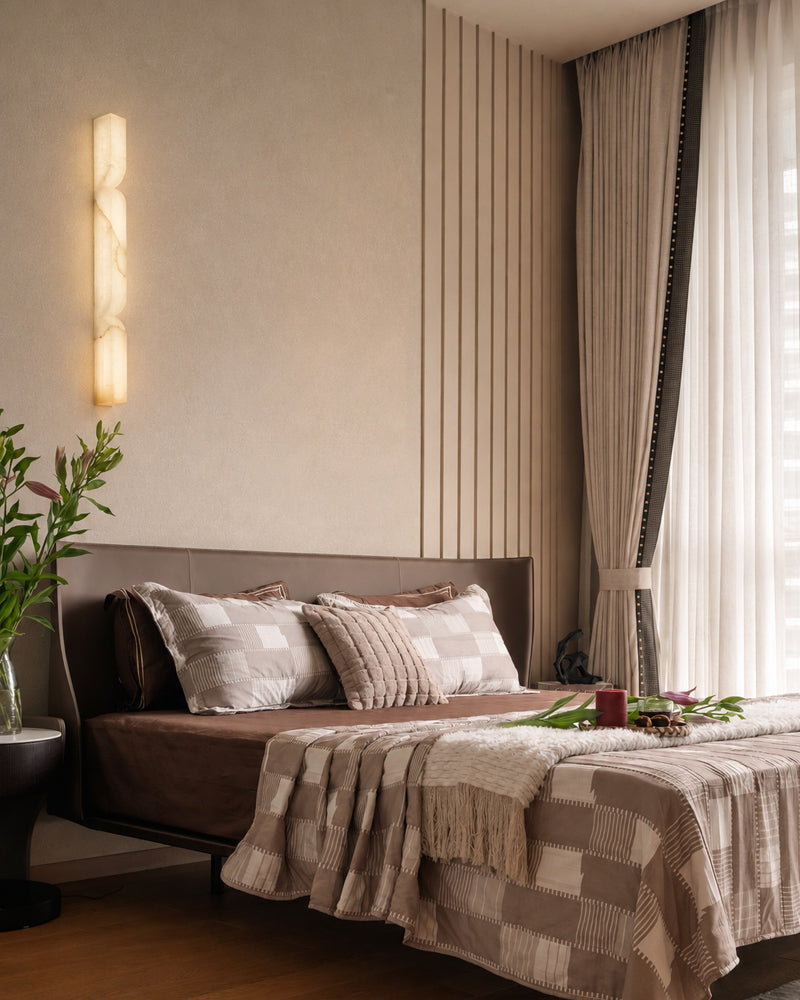 A modern bedroom featuring a bed dressed in brown and beige checkered bedding, a dark brown headboard, vertical wood paneling on the wall, a glowing vertical wall sconce, and sheer curtains by a window.