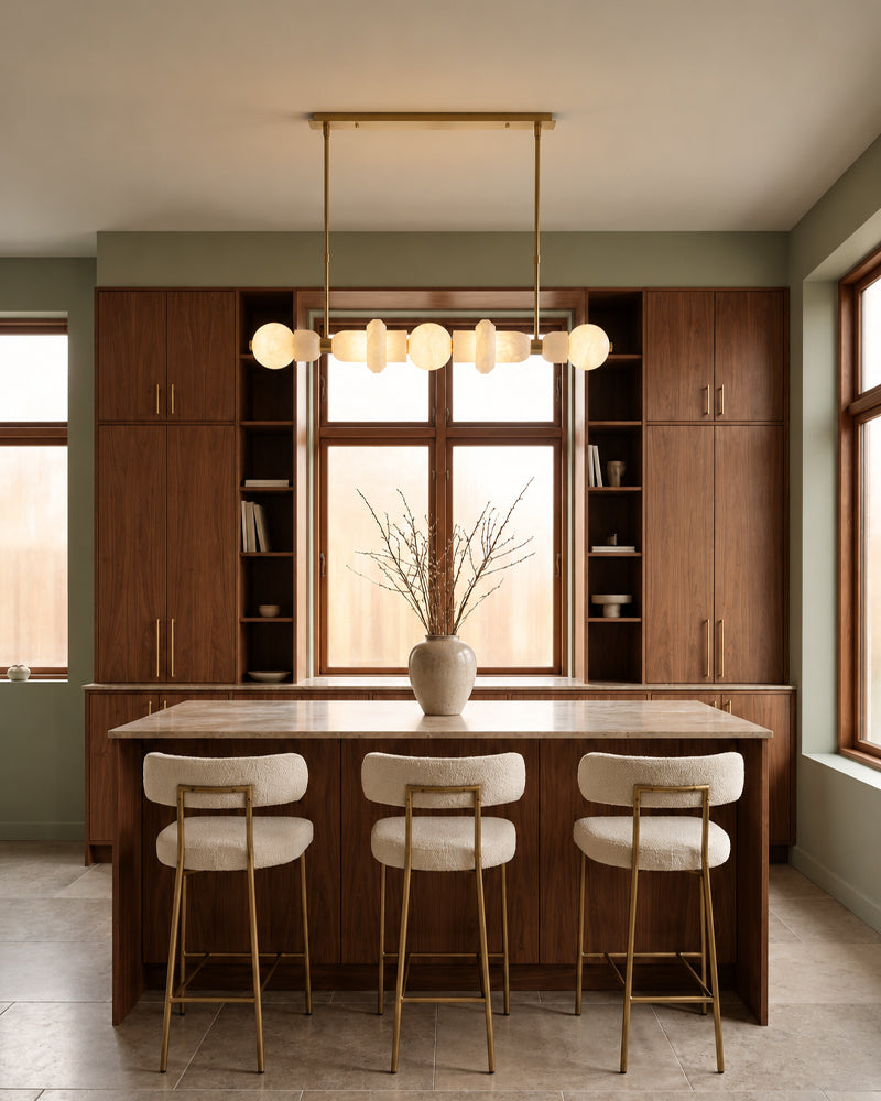 A modern kitchen island with a wood-look countertop and three upholstered bar stools with gold legs, set in a room with olive green walls, dark wood cabinetry, and a brass pendant light.